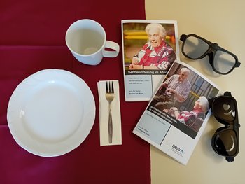 Ein weißer Teller und eine weiße Tasse stehen auf einer dunklen Tischdecke. Daneben liegen Broschüren und Simulationsbrillen.